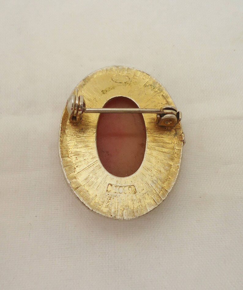 May include: Gold tone oval brooch with a pink cameo set in the center. The back of the brooch has a pin and a clasp. The brooch is marked "MCO."