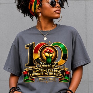 K&ouml;nnte beinhalten: Graues T-Shirt mit farbenfroher Grafik zum 10-j&auml;hrigen Jubil&auml;um. Das Design beinhaltet eine geballte Faust, den Text "Years of Honoring the Past, Empowering the Future" und die Jahreszahlen 1926-2026. Das T-Shirt hat einen Rundhalsausschnitt und kurze &Auml;rmel.