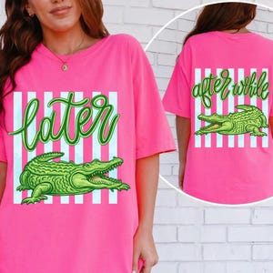 May include: Bright pink t-shirts with a green alligator graphic. The front of the shirt says "Later" in green script, and the back says "after while" in green script. White vertical stripes are behind the text and alligator.