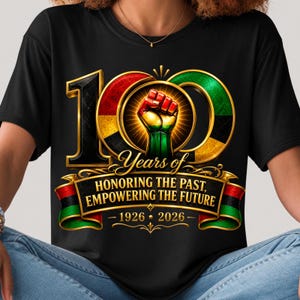 K&ouml;nnte beinhalten: Schwarzes T-Shirt mit einer goldumrandeten "100" Grafik, mit roten, gr&uuml;nen und schwarzen Akzenten. Das Design beinhaltet eine geballte Faust und den Text "Years of Honoring the Past, Empowering the Future 1926-2026."