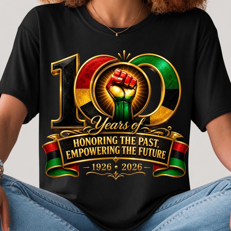K&ouml;nnte beinhalten: Schwarzes T-Shirt mit einer goldumrandeten "100" Grafik, mit roten, gr&uuml;nen und schwarzen Akzenten. Das Design beinhaltet eine geballte Faust und den Text "Years of Honoring the Past, Empowering the Future 1926-2026."