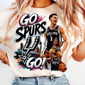 May include: A cream-colored t-shirt featuring a graphic print with the text GO SPURS GO!, a basketball player in a black jersey, a cartoon coyote mascot holding a basketball, and a stylized city skyline with the San Antonio Spurs logo.