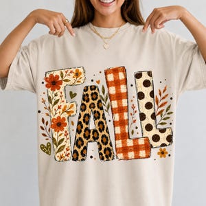 May include: A beige short-sleeve t-shirt featuring the word FALL printed in large letters. Each letter has a distinct pattern: the F has orange flowers, the A has a leopard print, the L has an orange plaid, and the L has brown polka dots.