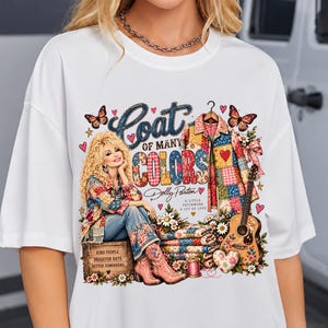 Può includere: Una t-shirt bianca con una stampa grafica di Dolly Parton seduta su una cassa di legno, un cappotto patchwork, una chitarra e farfalle. Il testo recita "Coat of Many Colors", "Dolly Parton", "A Little Patchwork A Lot of Love" e "Kind People Brighter Days Better Tomorrows".