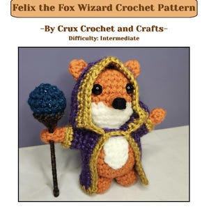 May include: A crochet fox wizard doll wearing a purple robe with gold trim and a hood, featuring a white belly and face, black eyes, and a dark nose. The fox holds a dark brown staff topped with a textured blue sphere. Text reads: Felix the Fox Wizard Crochet Pattern -By Crux Crochet and Crafts- Difficulty: Intermediate.