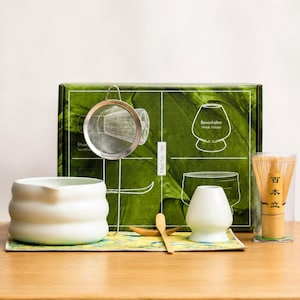 Peut inclure: Un ensemble de thé matcha avec un bol en céramique blanc, un support à fouet, un fouet en bambou et une passoire en acier inoxydable. L'ensemble comprend une cuillère en bois et est présenté sur un tapis décoratif, avec une boîte verte en arrière-plan.