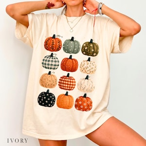 Puede incluir: Una camiseta de manga corta color marfil con un estampado gráfico de doce calabazas en varios colores y patrones, incluyendo diseños naranjas, verdes, negros, a cuadros, florales y punteados dispuestos en una cuadrícula.