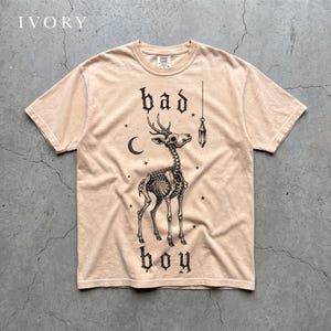 Puede incluir: Una camiseta de manga corta color marfil con un estampado gráfico negro de un esqueleto de ciervo, una luna creciente, estrellas y un colgante de cristal. El texto "bad boy" aparece impreso en una tipografía gótica sobre la prenda.