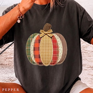 Puede incluir: Una camiseta de manga corta en color gris carbón con un gráfico grande de una calabaza hecha de varios parches de tela. El diseño incluye cuadros verdes, cuadros rojos, vichy amarillo y patrones florales con un pequeño lazo arriba.