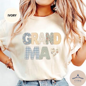 Puede incluir: Una camiseta de color marfil con la palabra GRANDMA en estilo patchwork con varios patrones, incluyendo cuadros vichy azules, cuadros amarillos y estampados florales. Un pequeño corazón floral se sitúa a la derecha de la palabra MA. Cuello redondo.