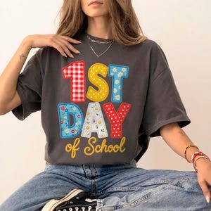 Puede incluir: Una camiseta de manga corta gris carbón con el texto 1ST DAY of School en letras de bloque coloridas y estampadas. El 1 es de cuadros rojos, el ST de lunares amarillos, DAY es de flores azules y margaritas blancas, y of School es amarillo.