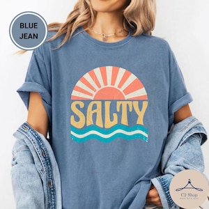 Puede incluir: Una camiseta de color azul vaquero con un estampado gráfico de un sol estilizado sobre olas y el texto SALTY en letras amarillas grandes. El diseño tiene una textura desgastada de estilo vintage. La camiseta tiene mangas cortas y cuello redondo.