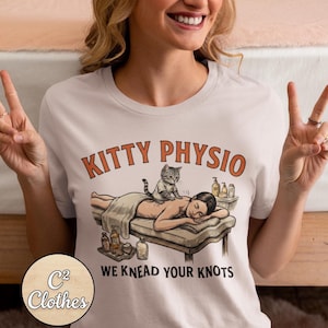 Puede incluir: Camiseta de color claro con la inscripción naranja "KITTY PHYSIO" sobre una ilustración de un gato masajeando a una persona. El texto "WE KNEAD YOUR KNOTS" está debajo. El logotipo de "C2 Clothes" es visible en la esquina inferior izquierda.