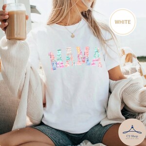 May include: White t-shirt with the word "MAMA" in colourful, patterned letters. The shirt has short sleeves and a crew neck. The word "WHITE" is in a white circle in the upper right corner.