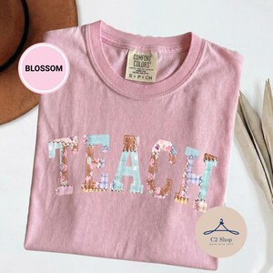 Puede incluir: Una camiseta de manga corta de color rosa claro con cuello redondo. La palabra TEACH está impresa en el pecho en letras grandes, cada una rellena con un patrón de patchwork diferente de flores, cuadros y telas lisas en marrón, azul y rosa.