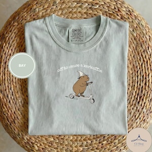 Puede incluir: Camiseta verde claro Comfort Colors con un gráfico de un carpincho montando un scooter y usando un sombrero de mago. El texto "off to cause a kerfuffle" está arriba. Talla S/P.