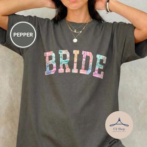 Puede incluir: Camiseta gris oscuro con la palabra "BRIDE" en letras coloridas y estampadas. La camiseta tiene mangas cortas y cuello redondo. La palabra "PEPPER" está en un círculo en el lado izquierdo.