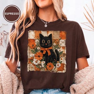 Puede incluir: Una camiseta marrón oscuro con un gráfico cuadrado de un gato negro con un lazo, rodeado de flores naranjas y blancas. El fondo del gráfico es un mosaico de texturas de tela naranja y beige. La palabra ESPRESSO aparece dentro de un círculo.