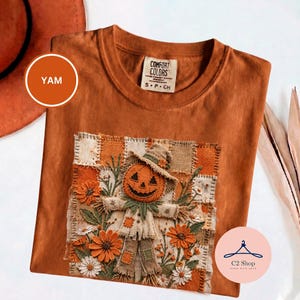 Puede incluir: Una camiseta de cuello redondo color óxido con etiqueta Comfort Colors y un parche cuadrado. El parche muestra un espantapájaros cosido con cabeza de calabaza, rodeado de flores bordadas en naranja y blanco sobre un fondo de tela a cuadros.