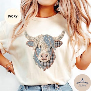 Puede incluir: Una camiseta de color marfil con un gráfico de una cabeza de vaca highland compuesto por varios parches de tela con patrones azules, blancos y negros, incluyendo estampados florales, de lunares y de cuadros vichy, con costuras visibles.