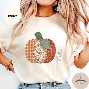 Puede incluir: Una camiseta de color marfil con un gráfico central de una calabaza compuesta por tres segmentos de estilo tela: uno con patrón de cuadros naranja, otro con estampado floral y otro con lunares blancos, con la palabra fall escrita en cursiva.