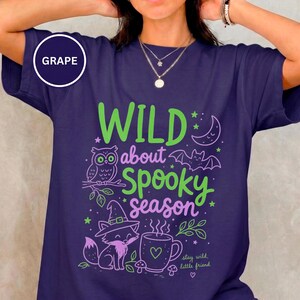 Puede incluir: Una camiseta color morado uva con un estampado gráfico que incluye el texto "WILD about SPOOKY season". El diseño muestra un búho verde, un murciélago morado, una luna creciente, un zorro con sombrero de bruja, una taza y pequeñas estrellas.