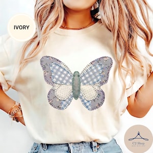 Puede incluir: Una camiseta color marfil con un gráfico de mariposa grande compuesto por varios parches de tela, incluyendo cuadros vichy azules y blancos, patrones florales y texturas de puntos, todo delineado con un efecto de bordado visible.