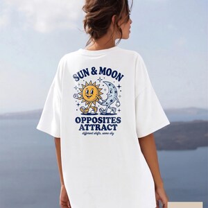 Puede incluir: Una camiseta blanca con un gráfico en la espalda que muestra un sol amarillo y una luna creciente azul tomados de la mano. El texto dice "SUN & MOON", "OPPOSITES ATTRACT" y "different shifts, same sky" en letras azules, rodeado de estrellas.
