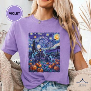 Puede incluir: Una camiseta de color morado claro con un estampado gráfico inspirado en un cielo estrellado sobre un cementerio. El diseño incluye calabazas naranjas, fantasmas blancos, una luna creciente amarilla y nubes azul oscuro con aves negras.