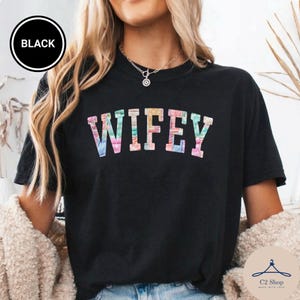 Puede incluir: Camiseta negra con la palabra "WIFEY" en letras coloridas de teñido anudado. La camiseta tiene cuello redondo y mangas cortas. Un círculo negro con la palabra "BLACK" está en la esquina superior izquierda.