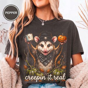 Puede incluir: Una camiseta gris carbón con un gráfico de una zarigüeya que lleva un gorro y un pañuelo a cuadros. La zarigüeya sostiene una taza y una calabaza. Otras calabazas, hojas de otoño y una telaraña rodean a la zarigüeya. El texto dice "creepin it real".