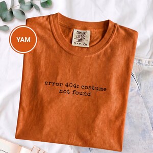 May include: A burnt orange crew neck t-shirt featuring the black text "error 404: costume not found" printed on the chest. The shirt has a Comfort Colors brand label at the inner neckline with size S P CH indicated. The fabric has a solid colour.