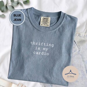 Puede incluir: Una camiseta Comfort Colors azul vaquero con el texto "thrifting is my cardio" en blanco. La camiseta es de talla pequeña y tiene cuello redondo. La camiseta está doblada sobre una superficie blanca.