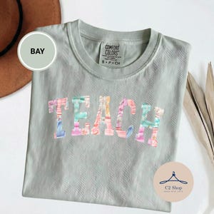 Puede incluir: Una camiseta verde salvia con la palabra "TEACH" en letras de patchwork coloridas. La camiseta tiene cuello redondo y mangas cortas. La etiqueta de la marca Comfort Colors es visible en la parte superior central de la camiseta.