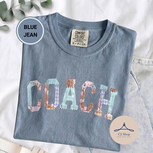 Puede incluir: Una camiseta color azul jean con cuello redondo y mangas cortas. El frente presenta la palabra COACH en letras grandes, cada una rellena con un patrón de patchwork diferente de telas florales y de cuadros en tonos azul, marrón y rosa.