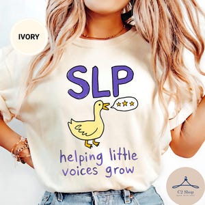 Puede incluir: Una camiseta de color marfil con un gráfico de un pato amarillo y un bocadillo de diálogo con tres estrellas naranjas. Sobre el pato aparece el texto morado "SLP" y debajo el texto morado "helping little voices grow" en letra informal.