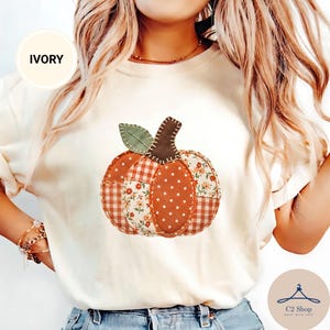 Puede incluir: Una camiseta de color marfil con un gráfico central de una calabaza hecha de varios patrones de tela, incluyendo cuadros vichy marrones y blancos, lunares naranjas y un estampado floral, con un tallo marrón oscuro y una hoja verde, todo con costuras.