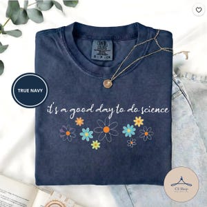 May include: A navy blue t-shirt with the phrase "it's a good day to do science" and colorful flower graphics. The shirt has a crew neck and short sleeves. A gold necklace with a pendant is draped across the shirt.