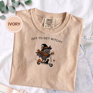 Puede incluir: Una camiseta color marfil con el gráfico de un murciélago con sombrero de bruja y bufanda de cuadros naranja, montado en un patinete con un café. Un gato negro pequeño y una calabaza aparecen cerca. El texto dice OFF TO GET WITCHY.