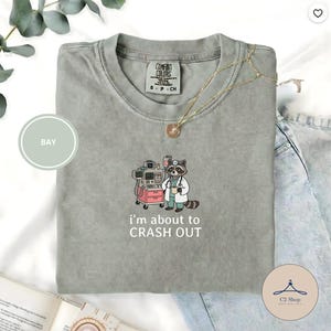 Puede incluir: Una camiseta Comfort Colors verde salvia con un bordado de un mapache de dibujos animados con equipo médico. El texto "I'm about to CRASH OUT" está debajo. También se ve un collar dorado y unos vaqueros de lavado claro.