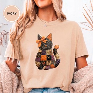 May include: An ivory short-sleeve t-shirt featuring a graphic of a sitting cat made of a patchwork quilt pattern. The cat has a large orange bow on its head and a tail that curves upward. The patchwork design includes various floral and celestial prints.