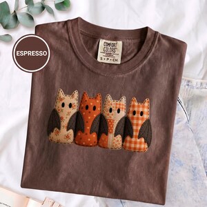 Puede incluir: Una camiseta color espresso con un gráfico de cuatro figuras de murciélago estilizadas. Cada murciélago tiene un patrón único, incluyendo diseños florales y de cuadros en tonos naranja y crema. La camiseta tiene una etiqueta Comfort Colors en el cuello.