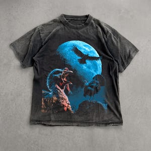 Puede incluir: Una camiseta de manga corta en color gris carbón con un estampado gráfico de dos pavos y un ave en vuelo frente a una gran luna llena de color azul brillante. La prenda tiene cuello redondo y un aspecto desgastado en el tejido.
