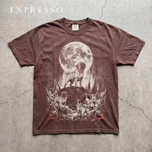 Puede incluir: Una camiseta marrón de manga corta con un estampado gráfico de tres carlinos en un bosque bajo una gran luna llena con murciélagos. El diseño incluye varias calabazas talladas con ojos de color naranja brillante y la palabra EXPRESSO.