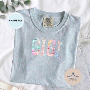 Puede incluir: Camiseta Comfort Colors azul claro con el nombre "GIGI" en letras de patchwork coloridas. La camiseta tiene cuello redondo y mangas cortas. La palabra "CHAMBRAY" está en un círculo azul claro a la izquierda.