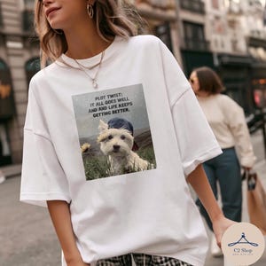 Puede incluir: Camiseta blanca con un gráfico de un perro pequeño en un campo y el texto "PLOT TWIST: IT ALL GOES WELL AND AND LIFE KEEPS GETTING BETTER." La camiseta tiene cuello redondo y mangas cortas.