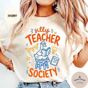 Puede incluir: Una camiseta de color marfil con un gráfico de un ganso con gafas, sentado en un taburete y leyendo un libro. El texto "silly TEACHER SOCIETY" aparece en naranja, acompañado de ilustraciones azules y amarillas de una bombilla, un cuaderno y banderines ABC.