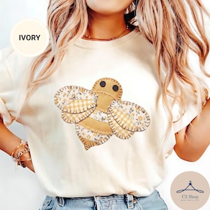 Puede incluir: Una camiseta de color marfil con un gráfico central de una abeja hecha de parches de tela. El diseño de la abeja incorpora cuadros vichy amarillos, patrones florales y secciones de tela beige sólida con bordes cosidos y dos ojos negros.