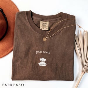 May include: A brown short-sleeved t-shirt with a crew neckline featuring the text "pie boss" and a graphic of a chef hat on a pie. The shirt has a Comfort Colors brand label on the inside collar and is displayed with a gold chain necklace.