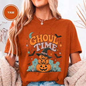 Puede incluir: Una camiseta de color naranja quemado con un estampado gráfico que incluye el texto GHOUL TIME en letras grandes de estilo retro. El diseño muestra tres calabazas apiladas con un sombrero de bruja, rodeadas de murciélagos, estrellas y velas.
